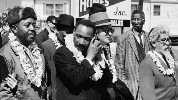 Martin Luther King, Jr. listening to a transistor radio in the front line of the third march from Selma to Montgomery, Alabama, to campaign for proper registration of black voters, March 23, 1965. Ralph Abernathy (second from left), Ralph Bunche (third from right) and Rabbi Abraham Joshua Heschel (far right) march with him.