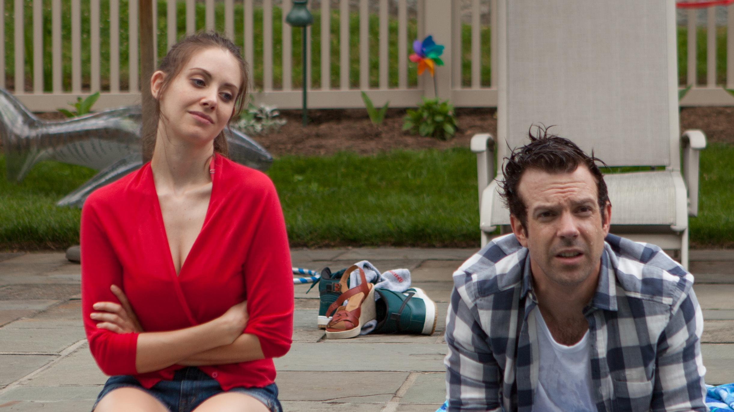 Image is from the film 'Sleeping With Other People' (2015). A man and woman sit next to each other beside a pool. The woman looks at the man, who is wearing a black plaid shirt. She is wearing a red cardigan and shorts.