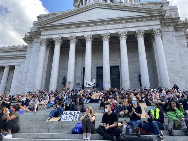 A crowd of people sits on the steps of the Capitol building in Olympia, with light gray tall pillars of the entrance at the top of the steps. 