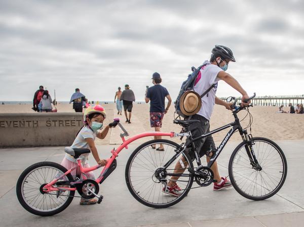 A girl and her father wear face masks while they push their bikes last summer in Hermosa Beach in the Los Angeles area. There aren't yet coronavirus vaccines approved in the U.S. for kids under 12 — which means they should keep masking, according to the CDC.