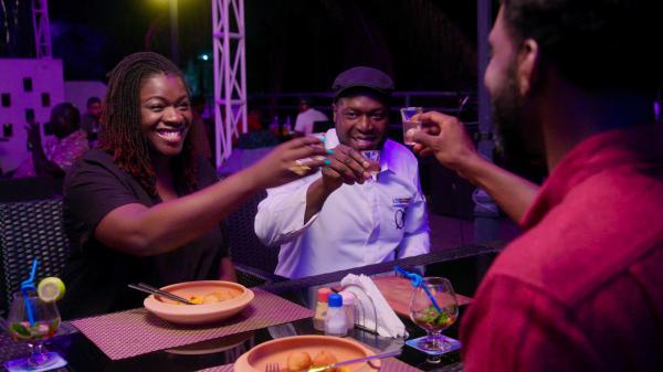 Benin's food scene is featured in the hit Netflix series <em>High on the Hog.</em> Pictured:<em> </em>Sedjro Ahouansou, center, head chef of Chill N Grill, raises a glass with local food blogger Karelle Vignon-Vullierme, left, and <em>High On The Hog </em>cohost Stephen Satterfield. Ahouansou is one of the chefs in the West African country who's reimagining traditional dishes.