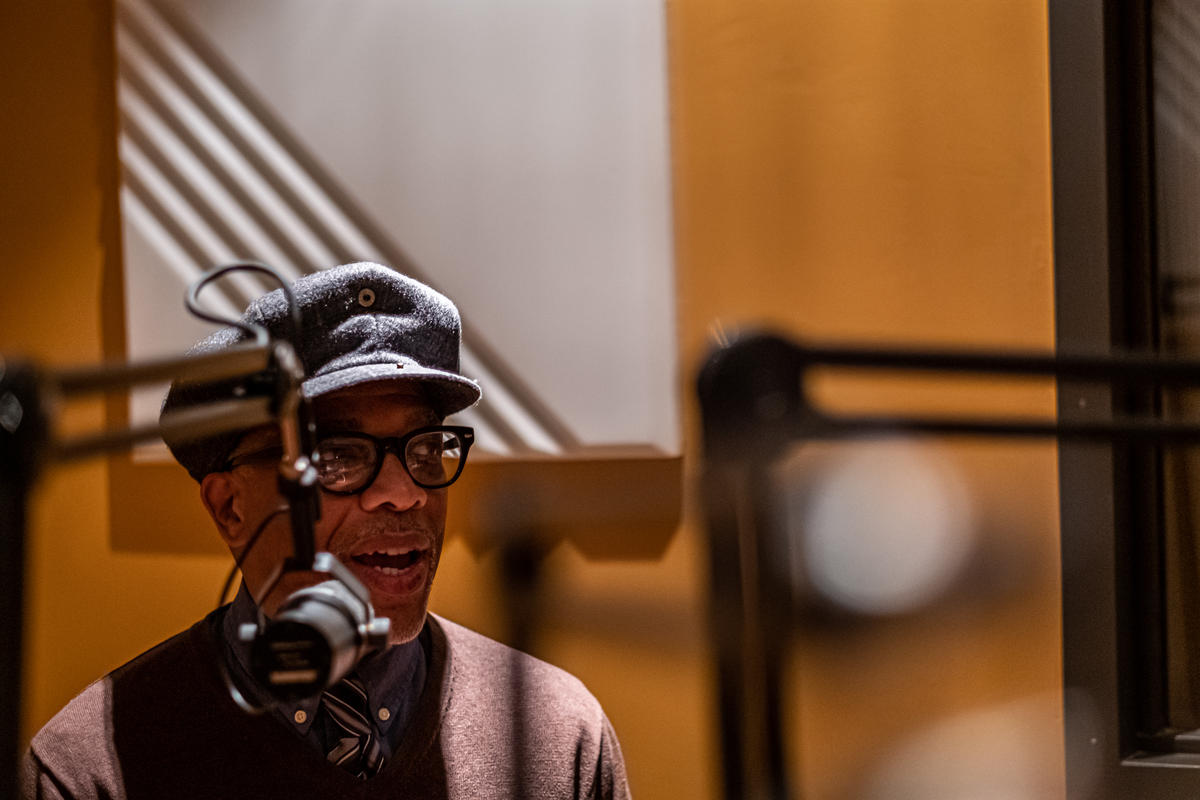 a man in a hat with glasses and a maroon shirt speaking into a microphone 